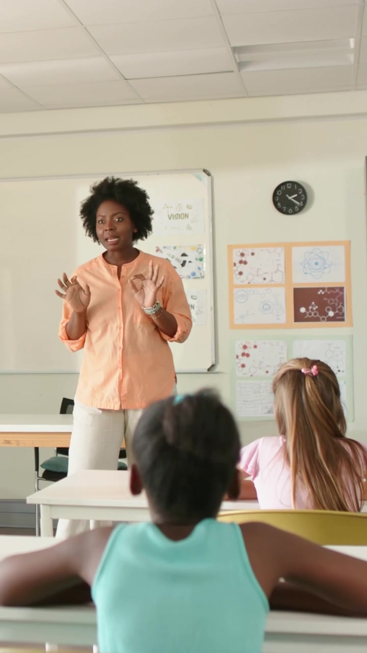 Gesturing teacher wearing peach blouse speaking to students at school, with board, posters, clock