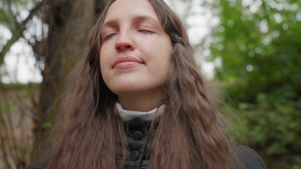 Giovane donna che cammina in un parco, lunghi capelli castani, cappotto nero, cielo nuvoloso, alberi, recinzione sullo sfondo, espressioni lente e pensierose, primo piano, respiro, aspirante poetessa che riflette in un momento contemplativo