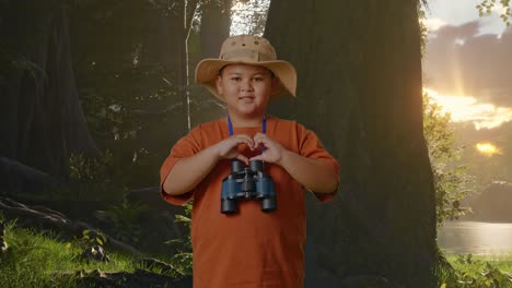 niño asiático con un sombrero y binoculares haciendo un gesto en forma de corazón mientras explora la naturaleza forestal. niño investigador examina algo, concepto de aventura de turismo de viaje