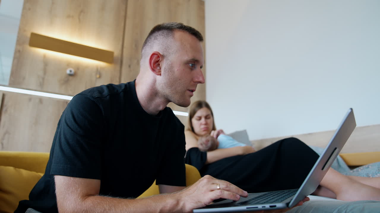 Focused Caucasian man has to work on his laptop being with his wife in maternity hospital. New-made mom takes care of the a baby at backdrop.