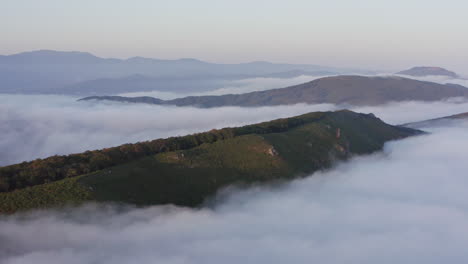 Mountain-ridge-with-trees-on-its-sides-surrounded-by-heavy-dense-clouds-of-fog-on-the-sunset