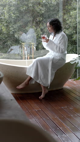 vertical video of biracial woman sitting on bathtub drinking tea at home, slow motion