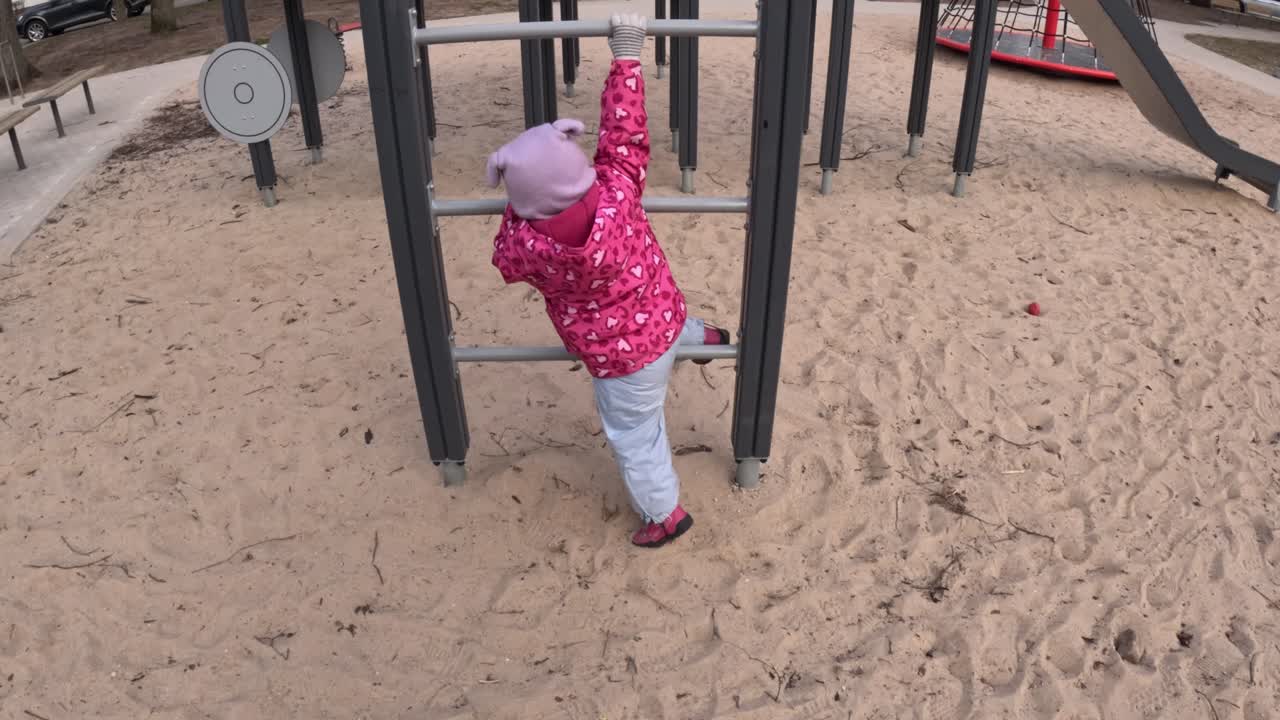 A toddler in a pink jacket struggles and enjoys the challenge of climbing on a playground, showcasing determination and playfulness.