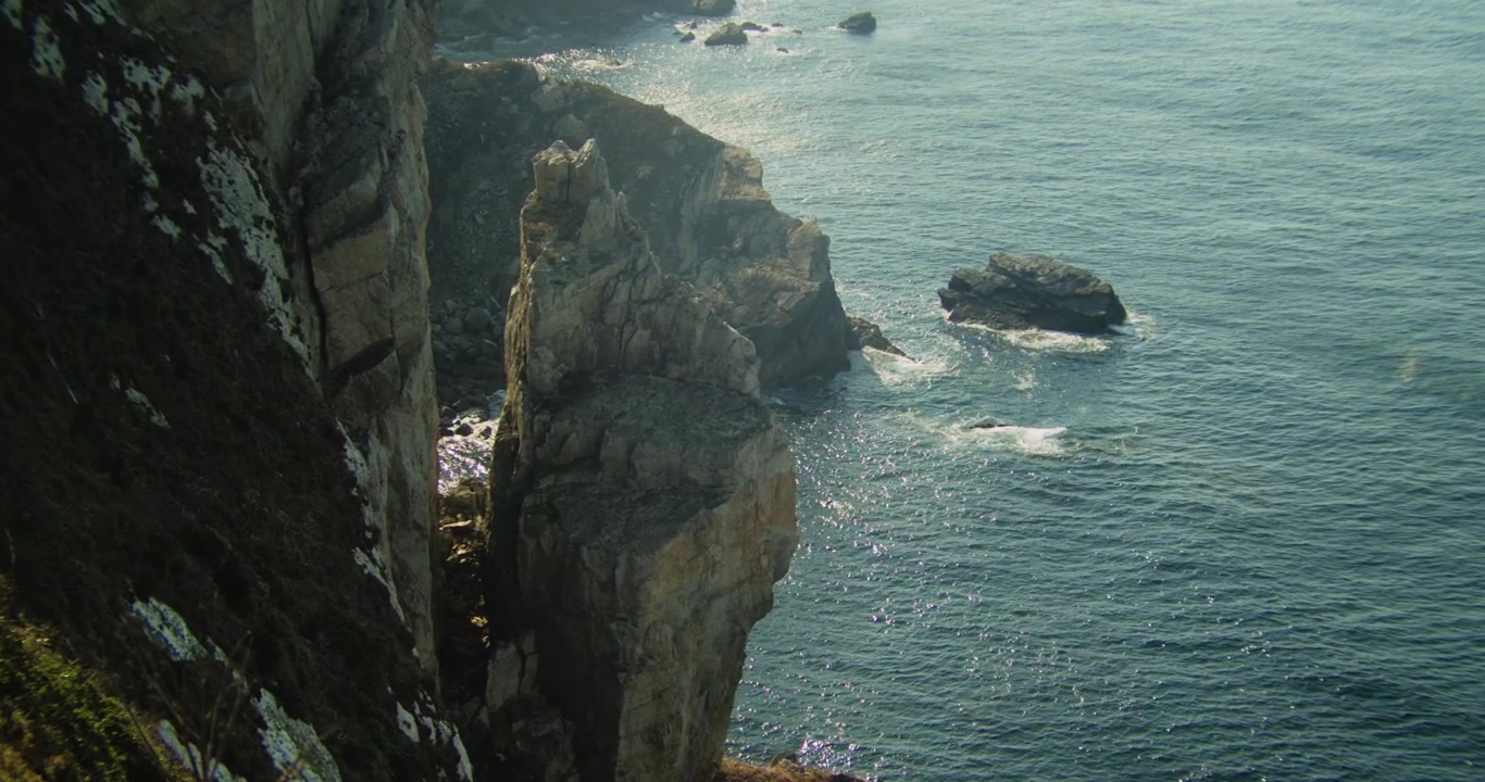 Handheld wide shot of tranquil Atlantic waves splashing against rugged Cabo Peñas cliffs, lush with native coastal plants on the wild coastline of Asturias under soft daylight.