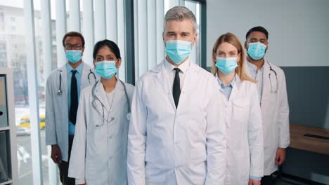 Portrait-Of-Serious-Experienced-Different-Mixed-Races-Healthcare-Workers-Doctors-And-Physicians-In-Medical-Masks-Posing-In-Clinic-Looking-At-Camera,-Quarantine-Concept,-Team-Colleagues