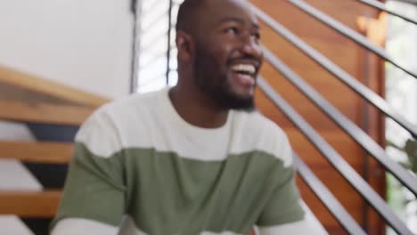 Video-of-happy-african-american-man-laughing,-sitting-on-stairs-at-home-holding-tablet