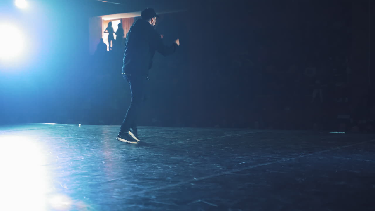 Talented young dancer dancing hip hop street dance on a stage in front of the spotlight. Back view of a man dancing to the audience in the club.