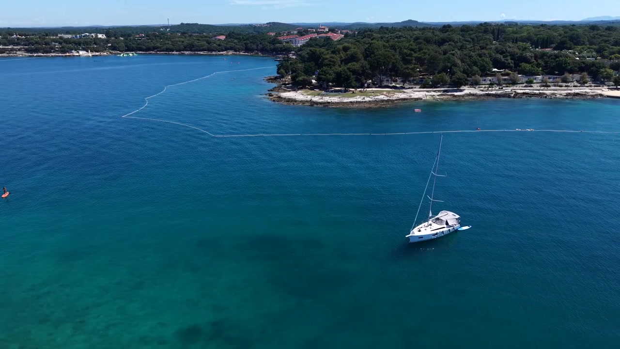 Penisola boscosa che si protende nel turchese Mare Adriatico lungo la costa istriana, Croazia, Europa, incorniciata da isole di pini, città costiera, barca a vela ancorata sotto un limpido cielo estivo blu. Ripresa aerea con drone in orbita.