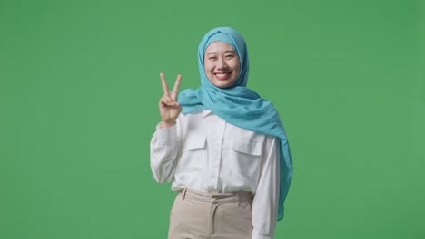 mujer musulmana sonriendo y mostrando un gesto de paz a la cámara mientras está de pie en el estudio de fondo de la pantalla verde