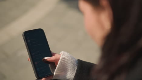 close-up of lady with short hair wearing black coat and white sweater scrolling on smartphone with focus on hand holding phone showing visible manicure