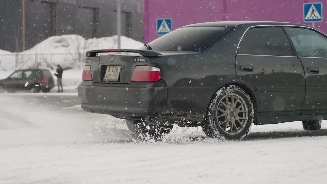 car performs controlled rotation on snowy road during winter with snow flying around spinning tires creating dynamic motion under cold cloudy sky with urban background including signs