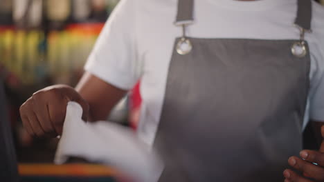 barman shakes rag against bar racks closeup. young waiter waves white cleaning cloth in restaurant. african american bartender cleans workplace