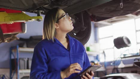 mecánica femenina usando una tableta digital de pie debajo de un coche en una estación de servicio de coches