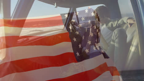 animation of american flag waving over biracial couple in car using smartphone by seaside