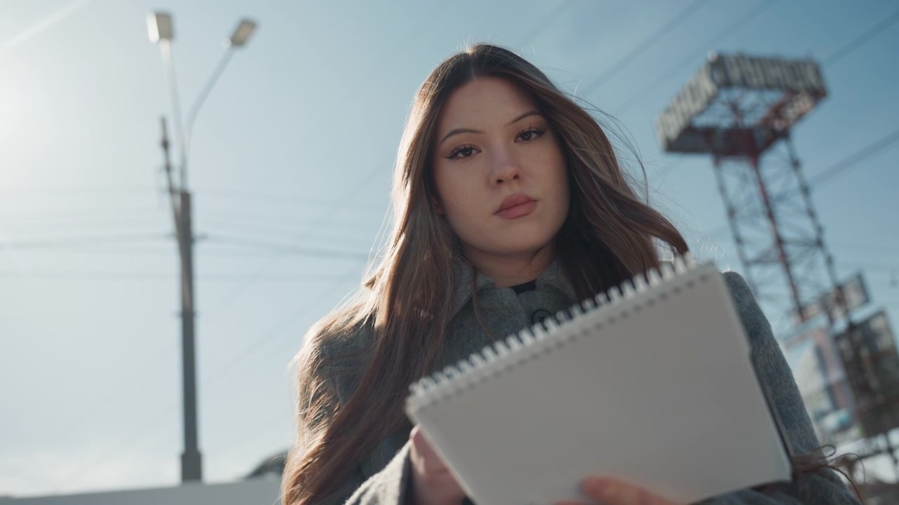 Bela escritora asiática escrevendo um diário sob o céu azul com caderno e caneta, expressão pensativa, casaco e cabelo iluminado por trás, postes telefônicos e torre industrial emoldurando a solidão urbana tranquila e o humor poético.