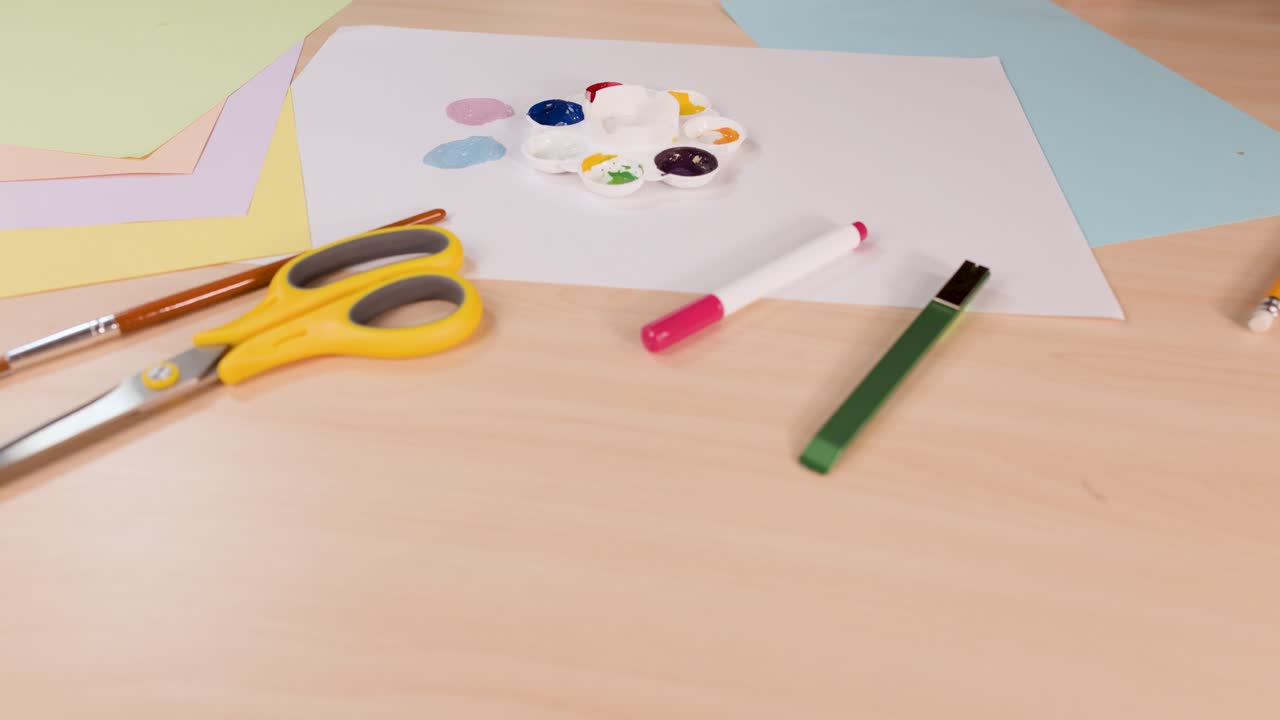 Hand mixes vibrant watercolor paints on palette, overhead view, soft lighting, art supplies visible