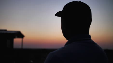 silhouetted man watching view of sunset with farmhouse in the background