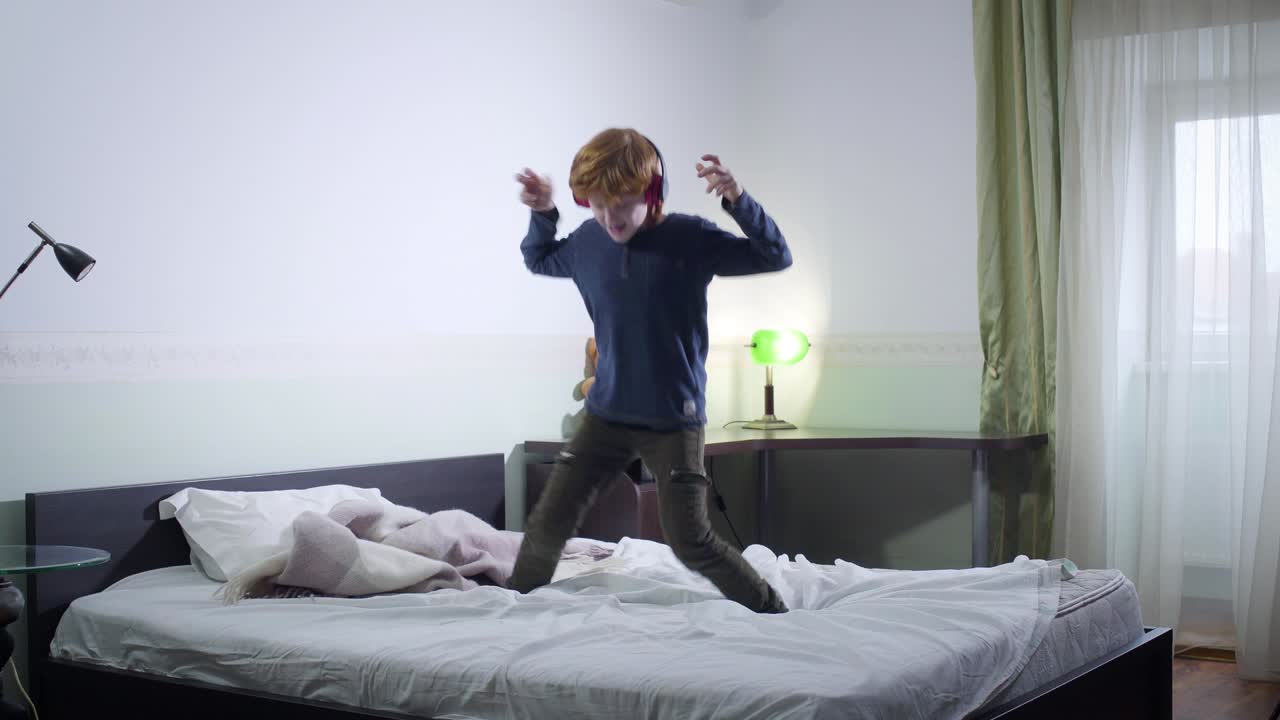 Active little redhead boy jumping on bed and listening to music. Cheerful Caucasian child having fun at home on weekends. Leisure, happiness, childhood.