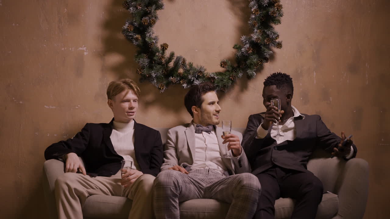 두 친구가 신년 파티에서 모바일을 사용하여 소파에 앉아 샴페인을 마시고 있고, 다른 친구가 앉아서 이야기를 시작합니다.