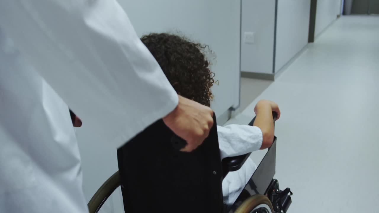 Close-up of Caucasian male doctor pushing pushing African american boy in wheelchair in hospital 4k