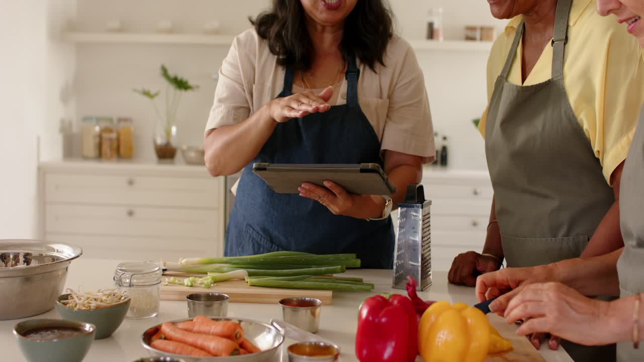 Amiche che seguono le istruzioni da un tablet sull'isola della cucina, tagliando i peperoni e preparando un pasto.