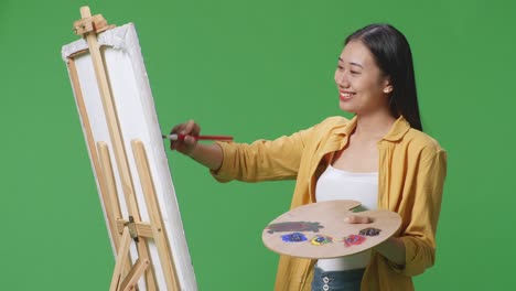 아시아 여성 예술가 가 녹색 스크린 스튜디오에서 오일 페인트와 으로 캔버스에 그림을 그리면서 카메라에 미소 짓고 있습니다.