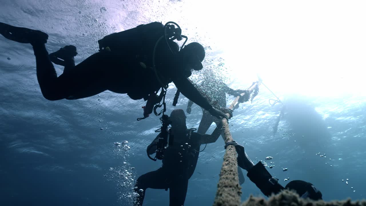 Scuba divers pause midwater while holding a descent line during a safety stop, silhouetted against bright surface light as streams of bubbles rise through the deep blue sea