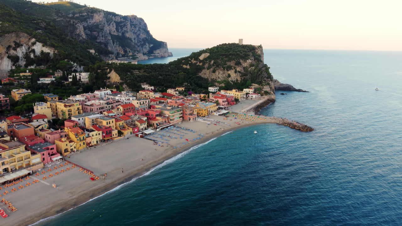 Ripresa aerea con drone che si allontana con un leggero movimento circolare, rivelando le case colorate di Varigotti, la spiaggia, le verdi colline circostanti e il mare al tramonto.