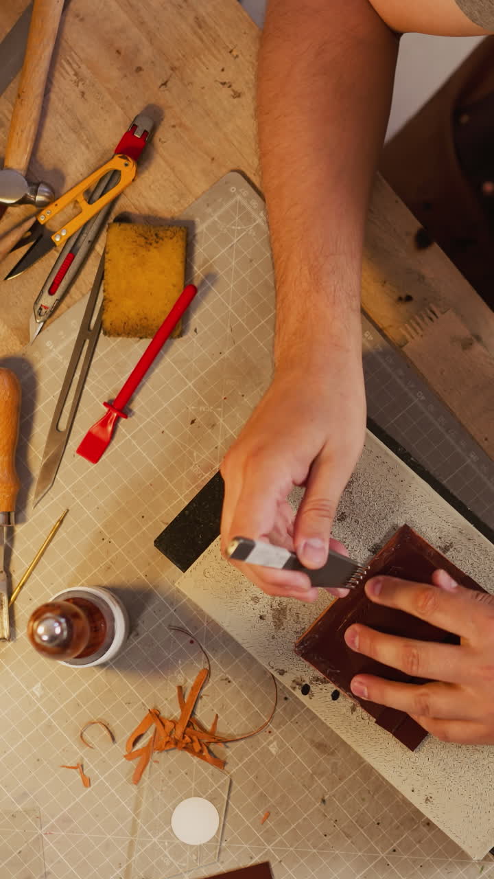 Worker pierces leather piece with awl and special round hammer at table in studio view from above. Tradition of handicraft leather items creation