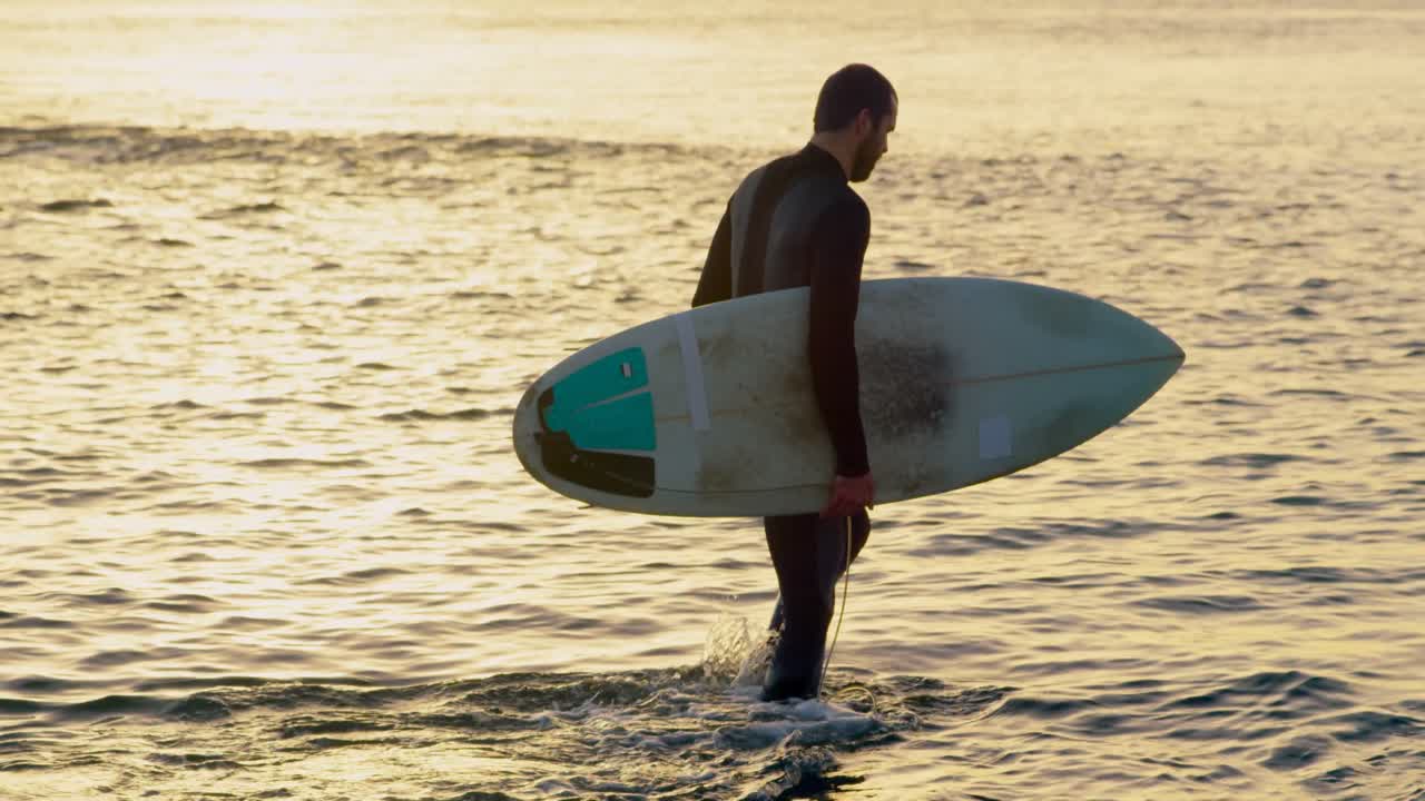 vista lateral de un surfista caucásico adulto con tabla de surf caminando en el mar durante la puesta de sol 4k
