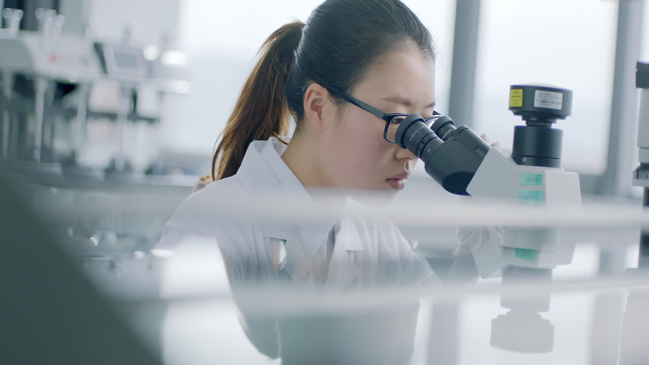 Smart female biologist or Scientist wearing a lab coat, looking into the microscope, Analyzes Petri Dish Sample. Specialists Working on Medicine, Biotechnology Research in Advanced Pharma Lab
