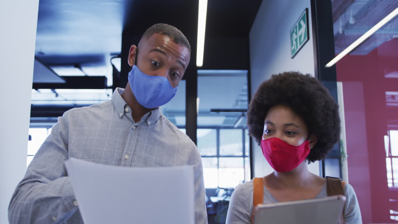 Diverse business people wearing face masks walking and using digital tablet in modern office