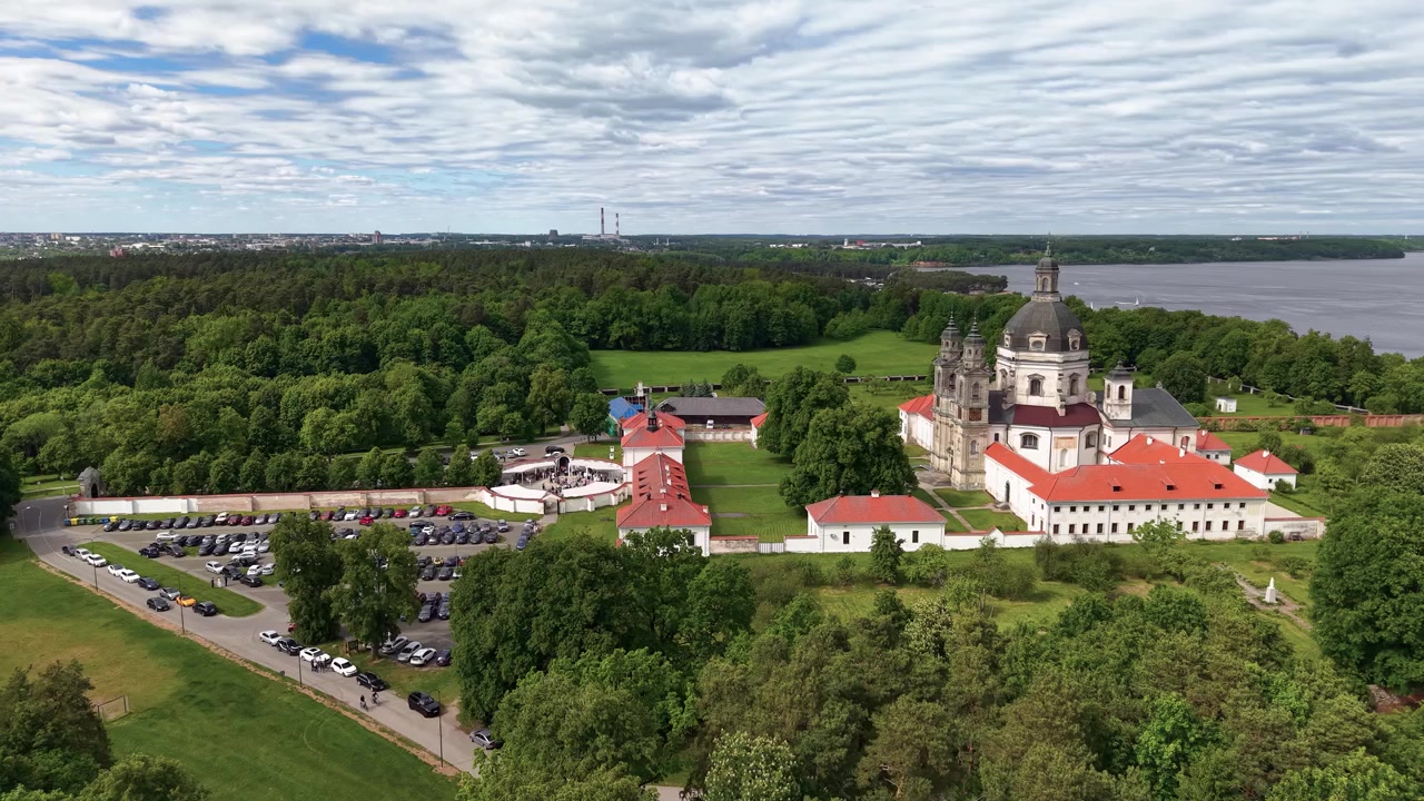 Una ripresa panoramica con drone rivela l'architettura barocca del Monastero di Pazaislis in mezzo a una lussureggiante foresta verde vicino a un lago a Kaunas, Lituania, sotto cieli nuvolosi e drammatici. Nessuna persona visibile.