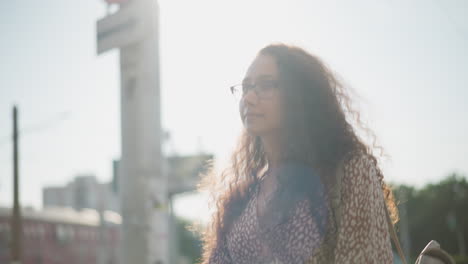 young woman in glasses walks in busy city with a soft smile, glancing sideways as cars pass by, against background of urban buildings and soft sunlight