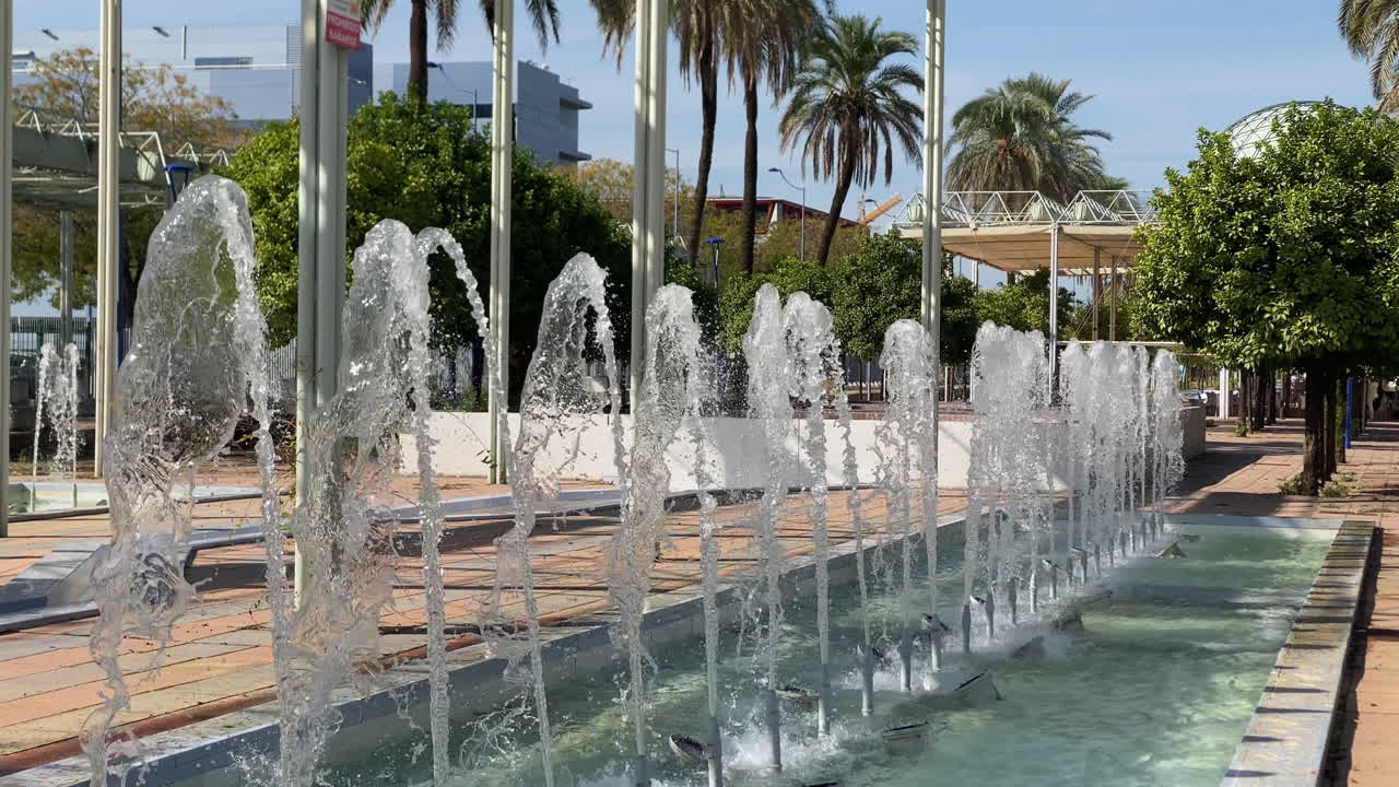 Unas fuentes modernas y una pérgola de metal enmarcan el parque fluvial Expo de Sevilla, con el gran pabellón esférico al fondo.
