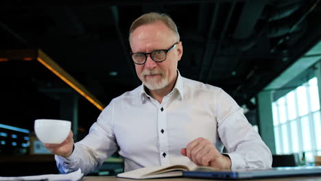 positive caucasian mature businessman arranging his working place at desk. man starting his workday from a cup of coffee enjoying the drink much. close up.
