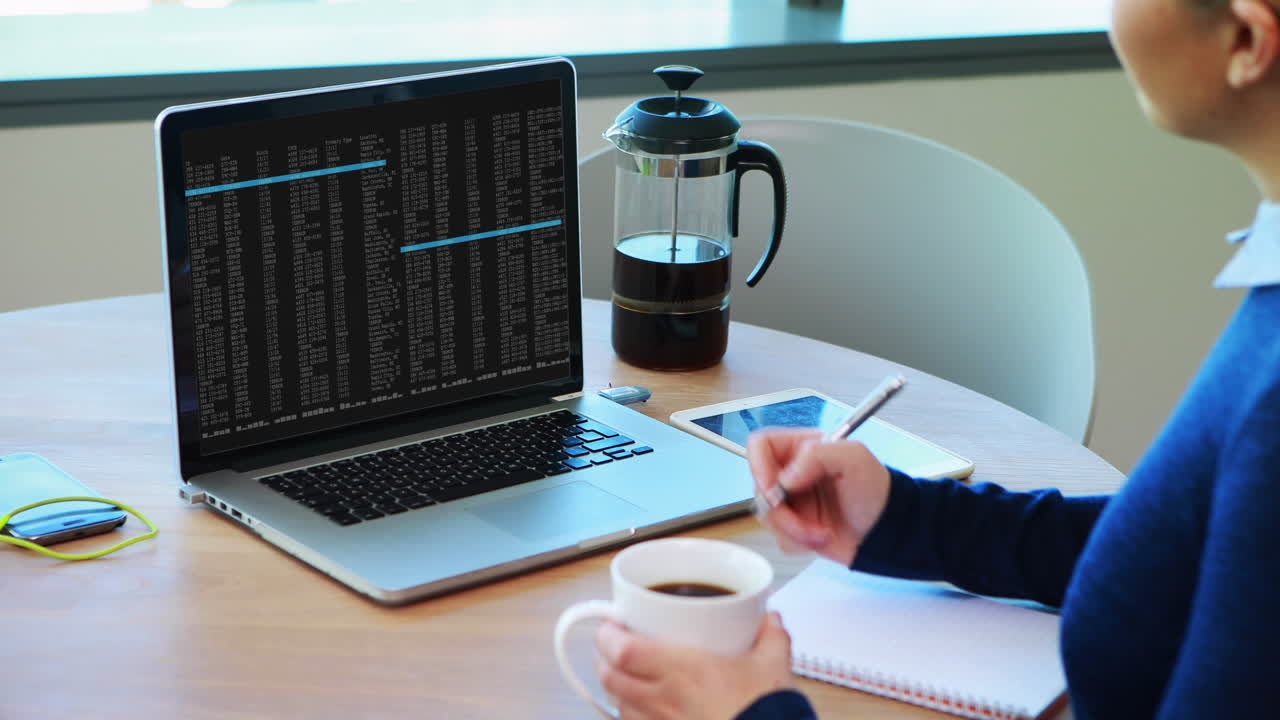 donna caucasica seduta a un tavolo a guardare l'elaborazione dei dati di codifica sullo schermo del portatile e a prendere appunti