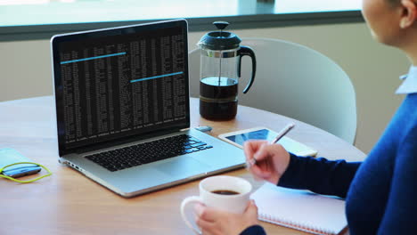 mujer caucásica sentada en una mesa viendo el procesamiento de datos de codificación en la pantalla de la computadora portátil y tomando notas