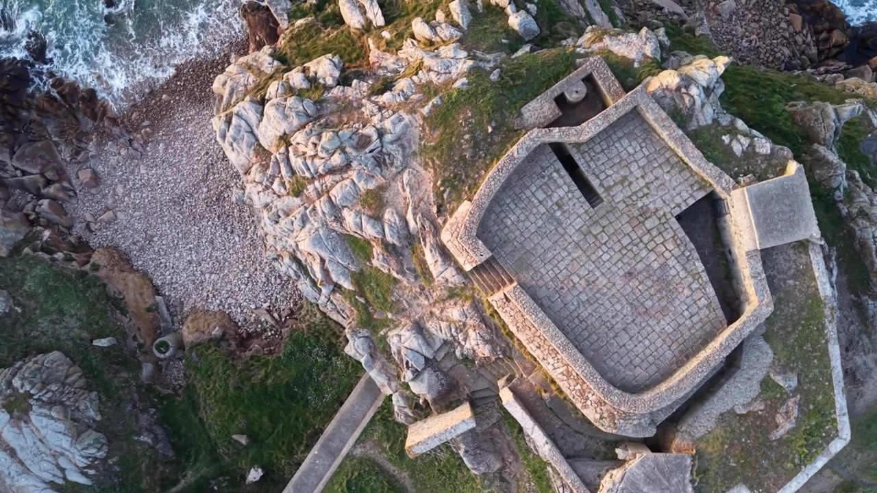 Grandes Rocques, Guernsey, Isole del Canale. Riprese aeree con drone che volteggia e scende su una piccola spiaggia con acqua cristallina alla base di un vecchio forte militare arroccato su rocce di granito al tramonto.