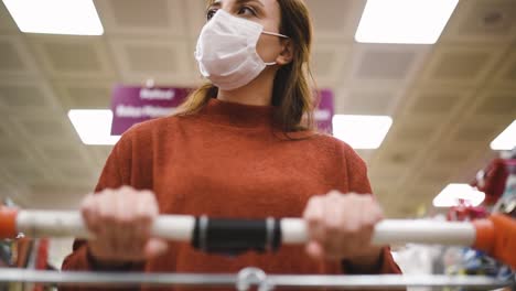 beautiful woman wearing protective disposable medical mask and fashionable clothes uses shopping cart while shopping in supermarket