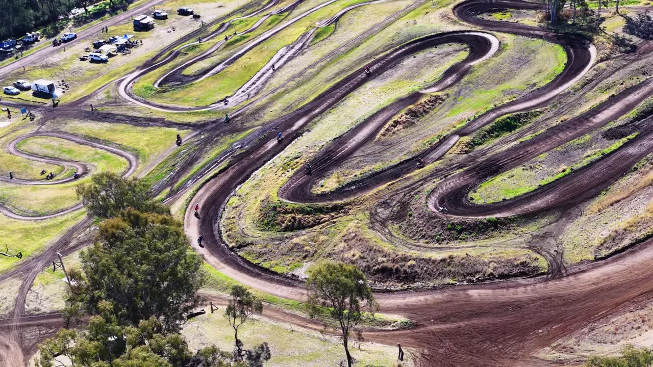 Drone captures winding dirt motocross track, parked vehicles, and trees under bright daylight conditions