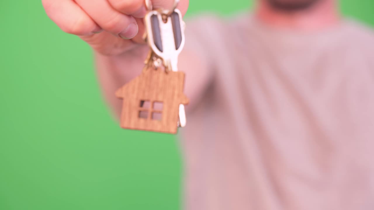 Male hand raises keys with a keychain in the shape of a house on a green background Chromakey