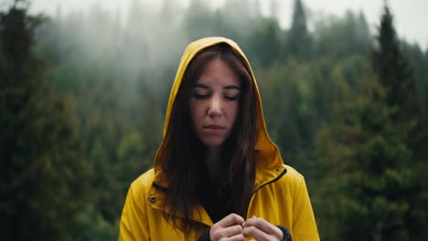 A-sad-girl-in-a-yellow-jacket-with-a-hood-stands-against-the-backdrop-of-a-coniferous-mountain-forest,-she-is-sad-and-looks-at-the-camera