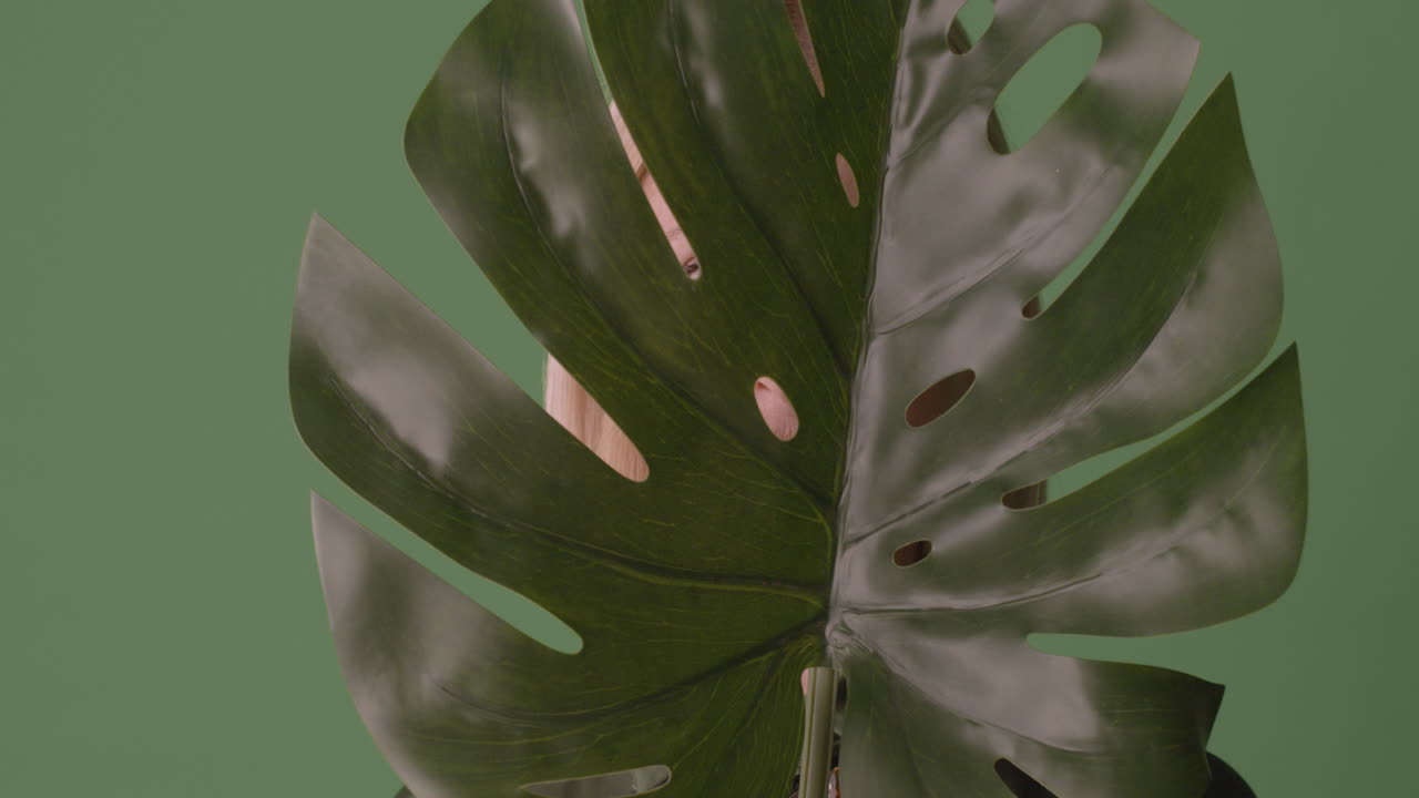 금발의 성숙한 여인은 큰 잎사귀로 얼굴을 가린 다음 웃는 얼굴을 보여주고 녹색 배경의 카메라를 바라본다