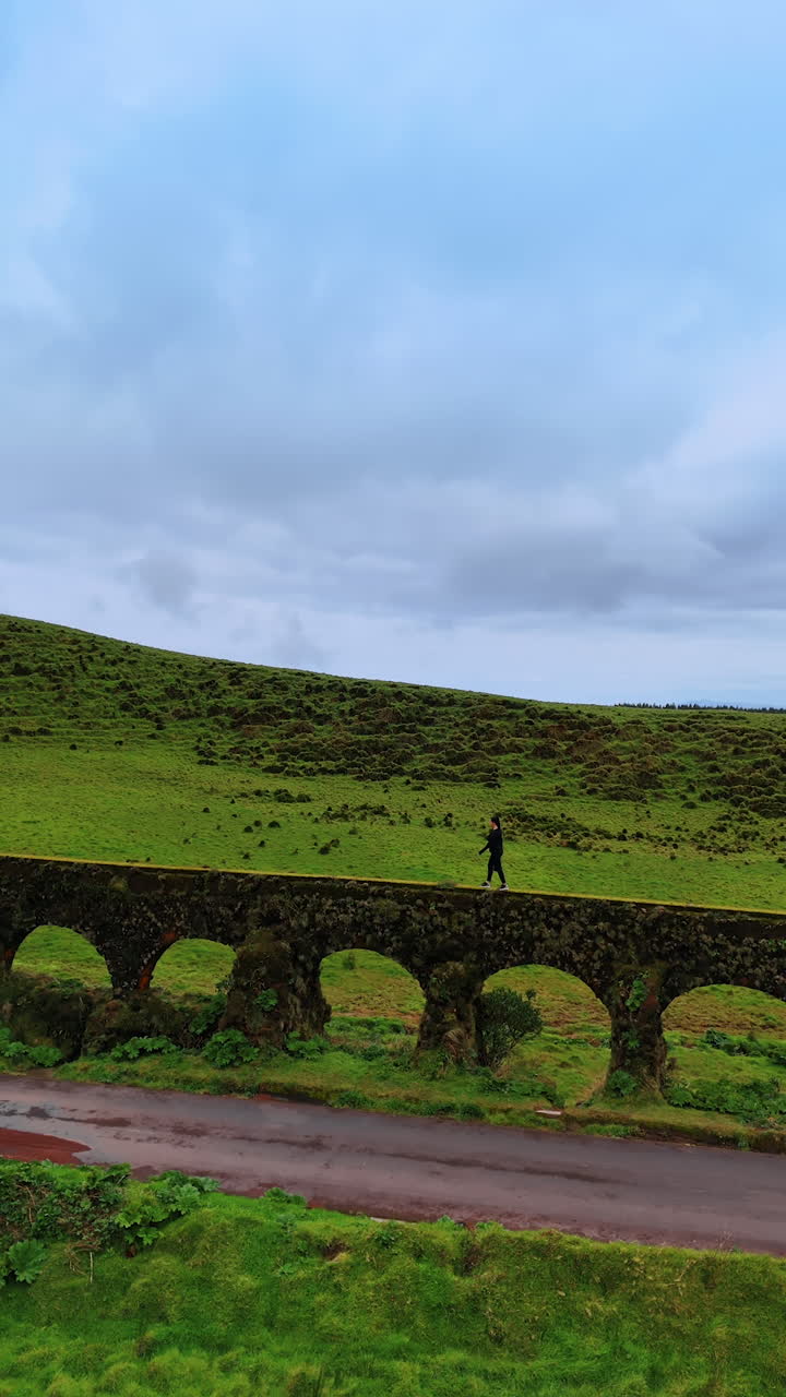 Woman walks by the viaduct along the asphalt road. Green valleys and hills at backdrop. Azorean Islands. Vertical video.