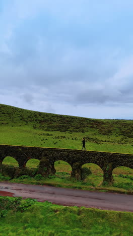 woman walks by the viaduct along the asphalt road. green valleys and hills at backdrop. azorean islands. vertical video.
