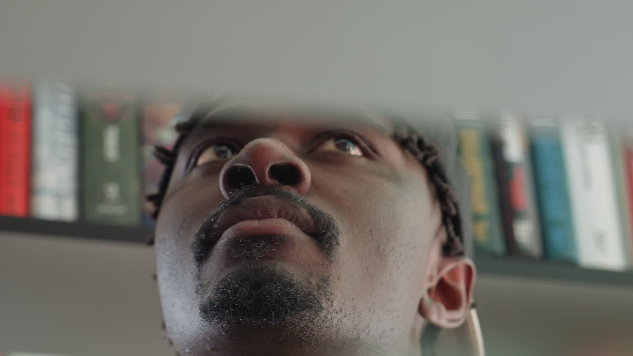 Extreme low angle close up of young man looking upward with focused eyes, face partially illuminated, set against blurred background of colorful books on shelves