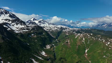 flying above kenai fjords national park with lush green mountains partly covered by snow under blue cloudy sky in alaska, usa