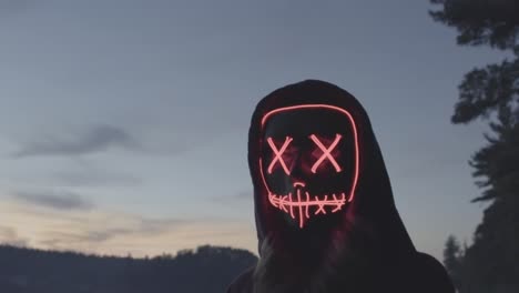 Close-up-of-mysterious-girl-wearing-red-glow-Halloween-mask-at-dusk-near-river-bank,-Slow-motion