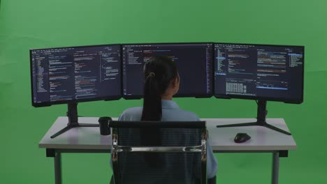 vista trasera de una desarrolladora asiática que tiene dolor de cabeza mientras escribe código con múltiples pantallas de computadora en el estudio de pantalla verde